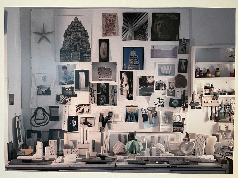 A photograph of Kim Lim's studio pinboard, displaying a collage of visual references, including photographs, sketches, and objects, alongside shelves containing tools and materials.